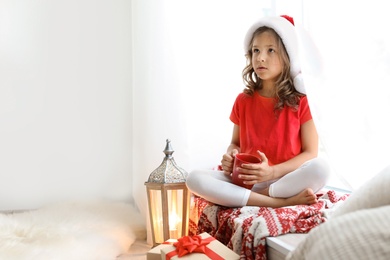 Photo of Cute little child in Santa hat with cup of cocoa sitting on windowsill at home. Christmas celebration