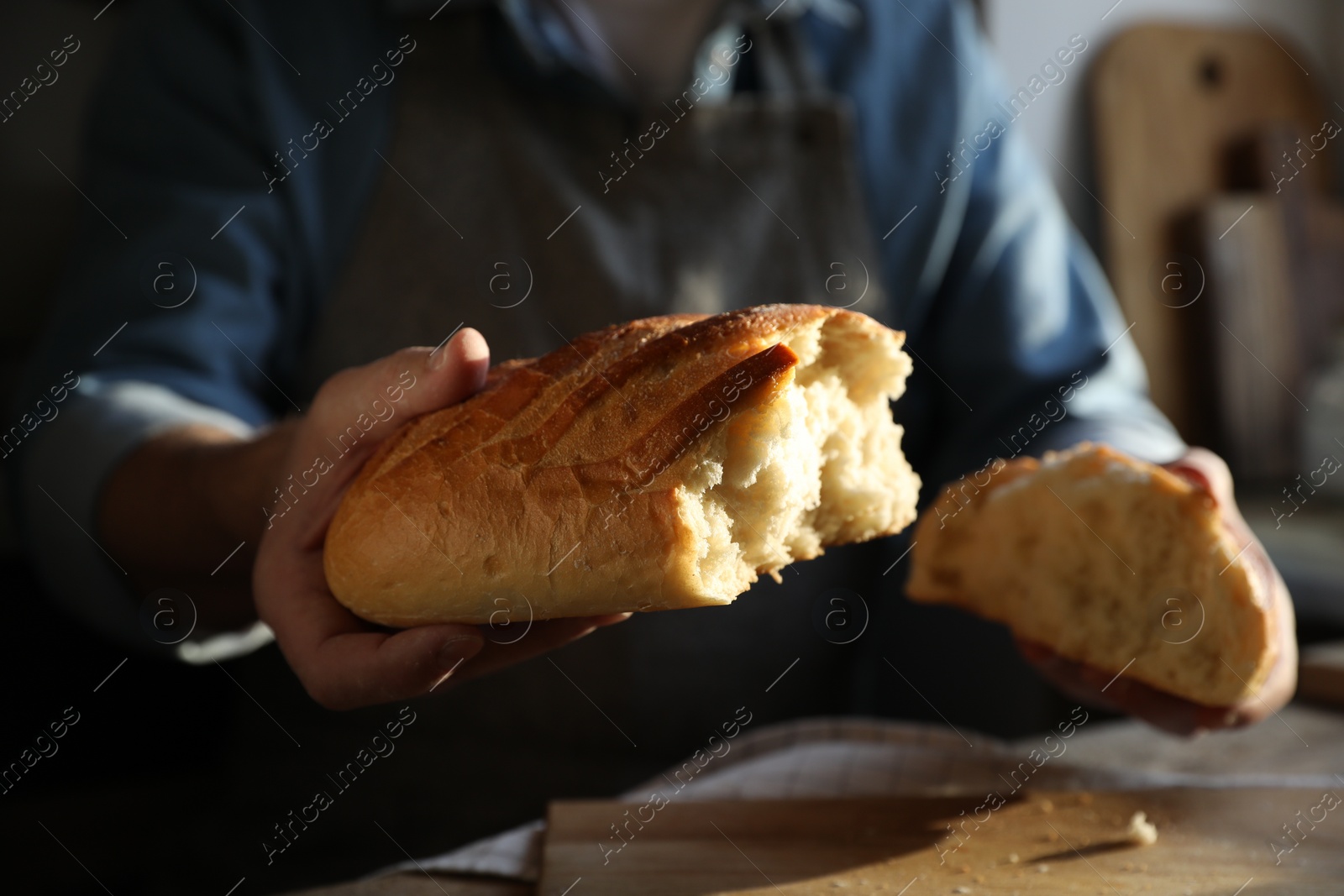 Photo of Man breaking loaf of fresh bread at wooden table indoors, closeup