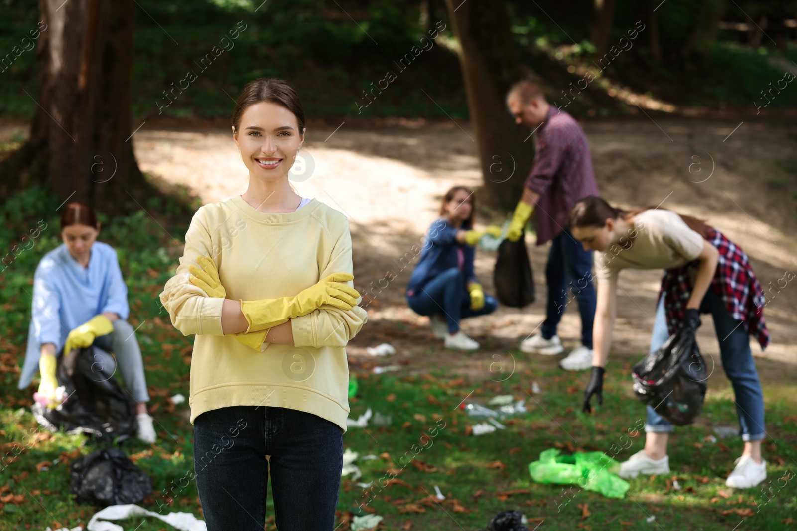 Photo of Young woman in gloves and group of people with plastic bags collecting garbage outdoors