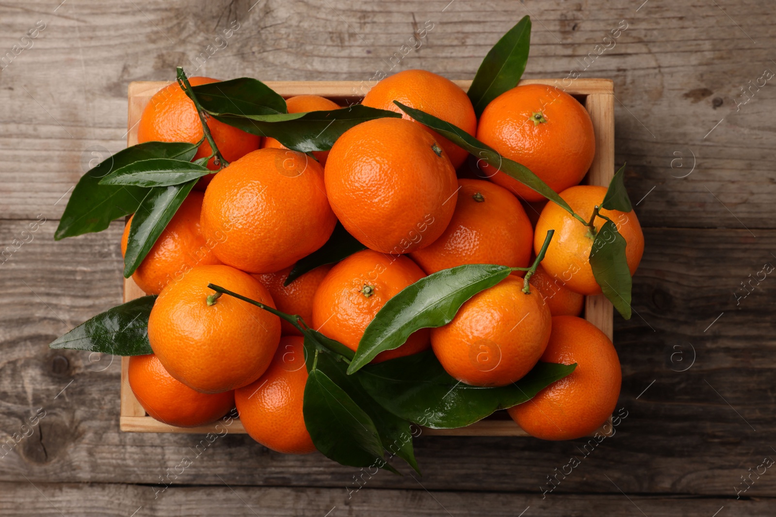 Photo of Delicious tangerines with leaves in crate on wooden table, top view