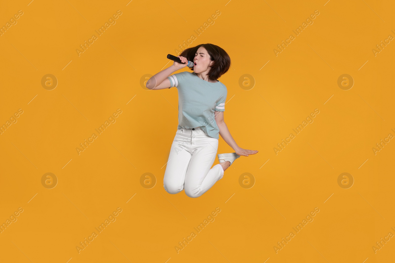 Photo of Beautiful young woman with microphone singing and jumping on yellow background