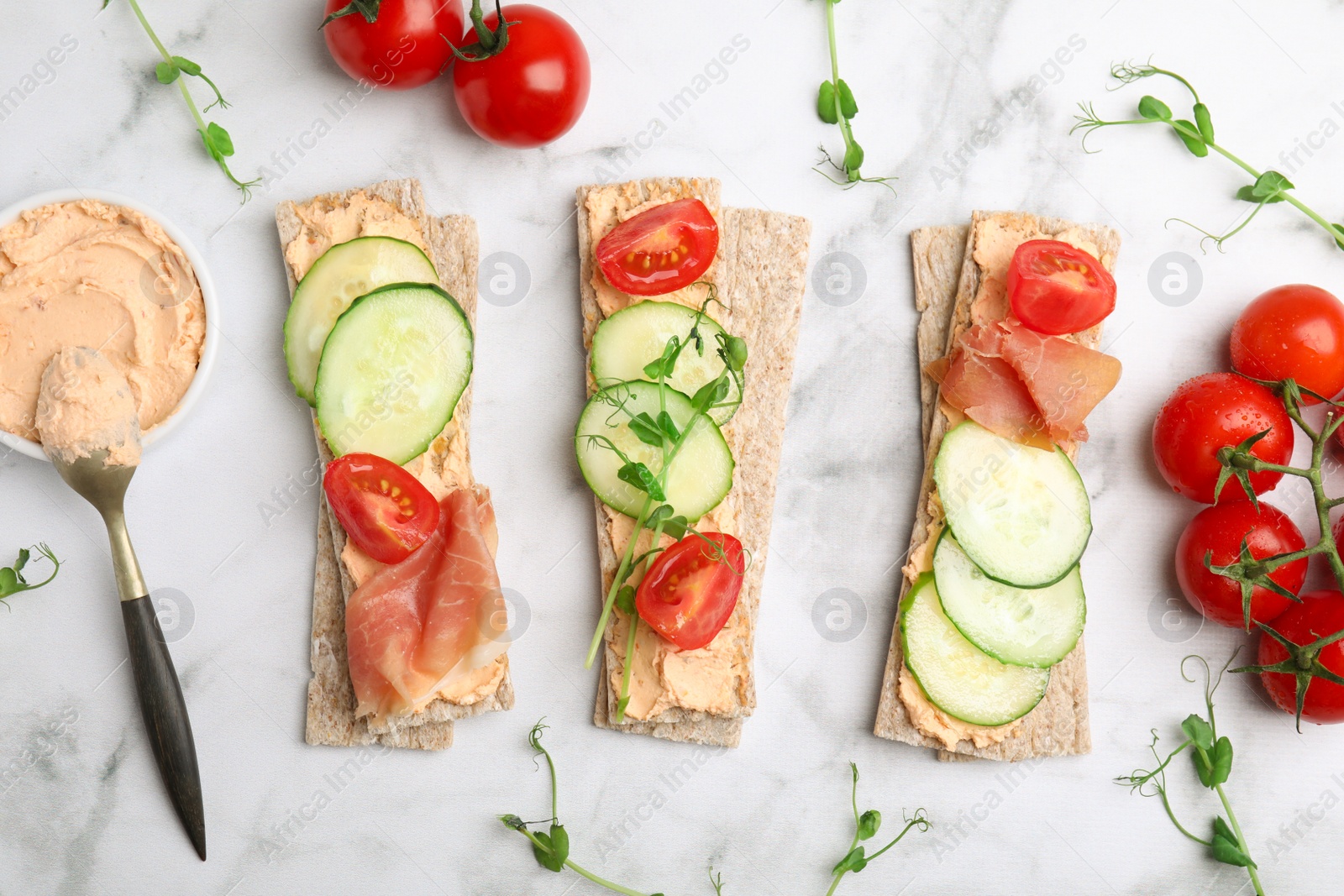 Photo of Tasty crispbreads with prosciutto, cream cheese and vegetables on white marble table, flat lay