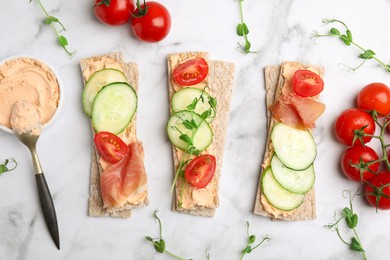Photo of Tasty crispbreads with prosciutto, cream cheese and vegetables on white marble table, flat lay