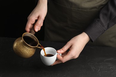 Photo of Turkish coffee. Woman pouring brewed beverage from cezve into cup at black table, closeup