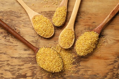 Spoons with raw bulgur on wooden table, flat lay