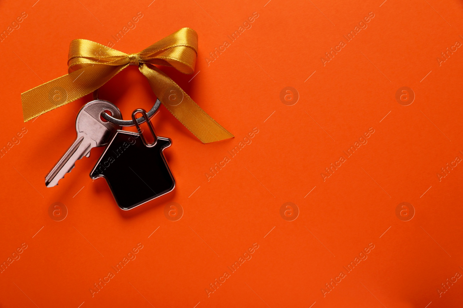 Photo of Key with trinket in shape of house and yellow bow on orange background, top view. Space for text. Housewarming party