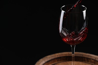 Pouring red wine into glass on wooden barrel against black background, closeup. Space for text