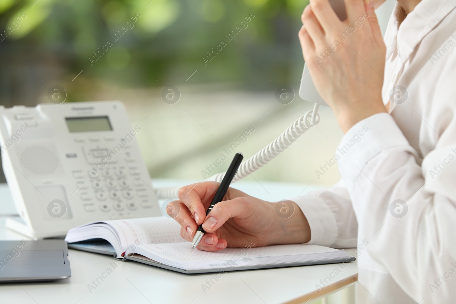 Photo of Assistant with telephone handset writing at white table against blurred green background, closeup