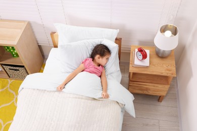 Little girl lying in bed and looking at alarm clock on bedside table indoors, above view