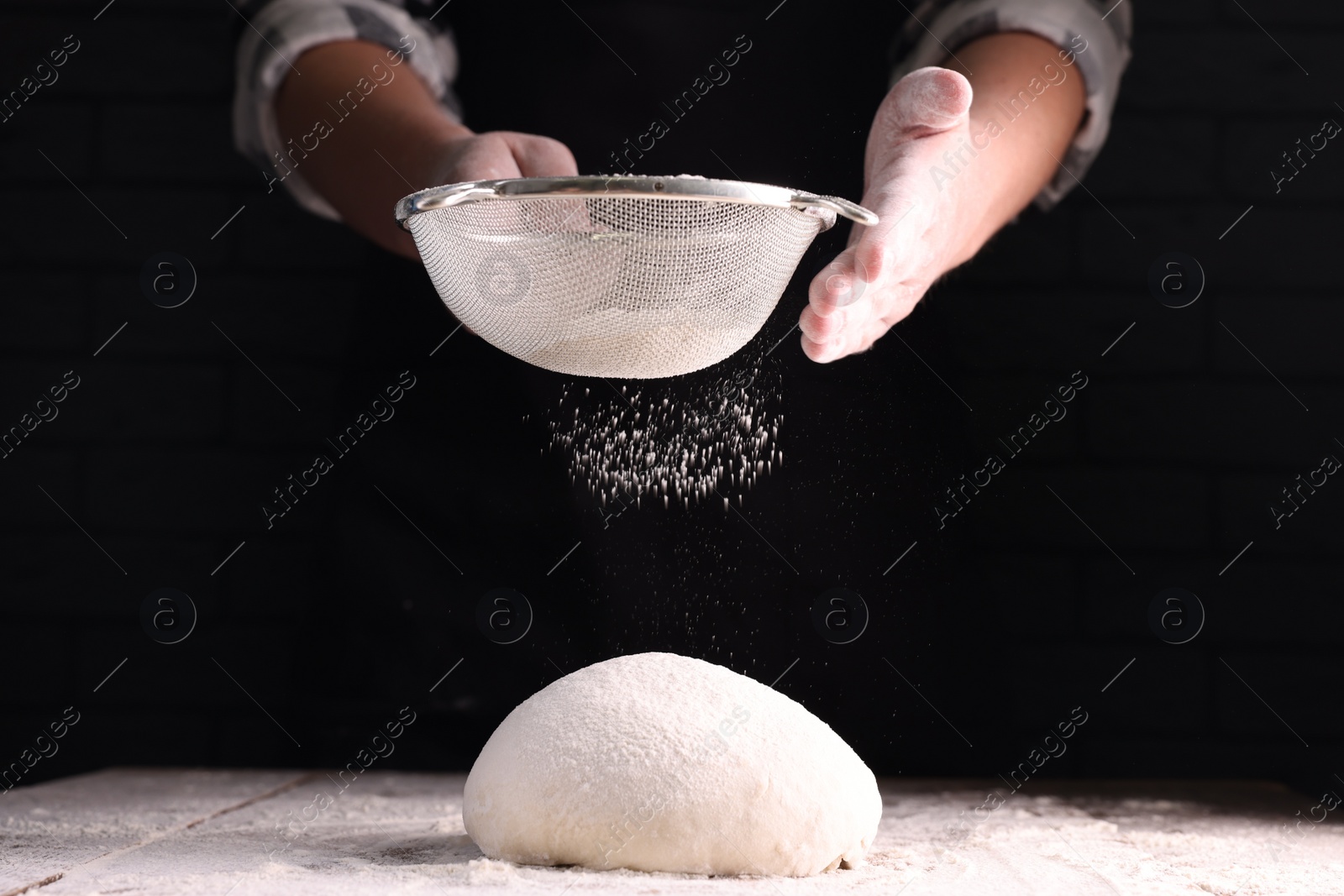 Photo of Man sprinkling flour over dough at wooden table on dark background, closeup