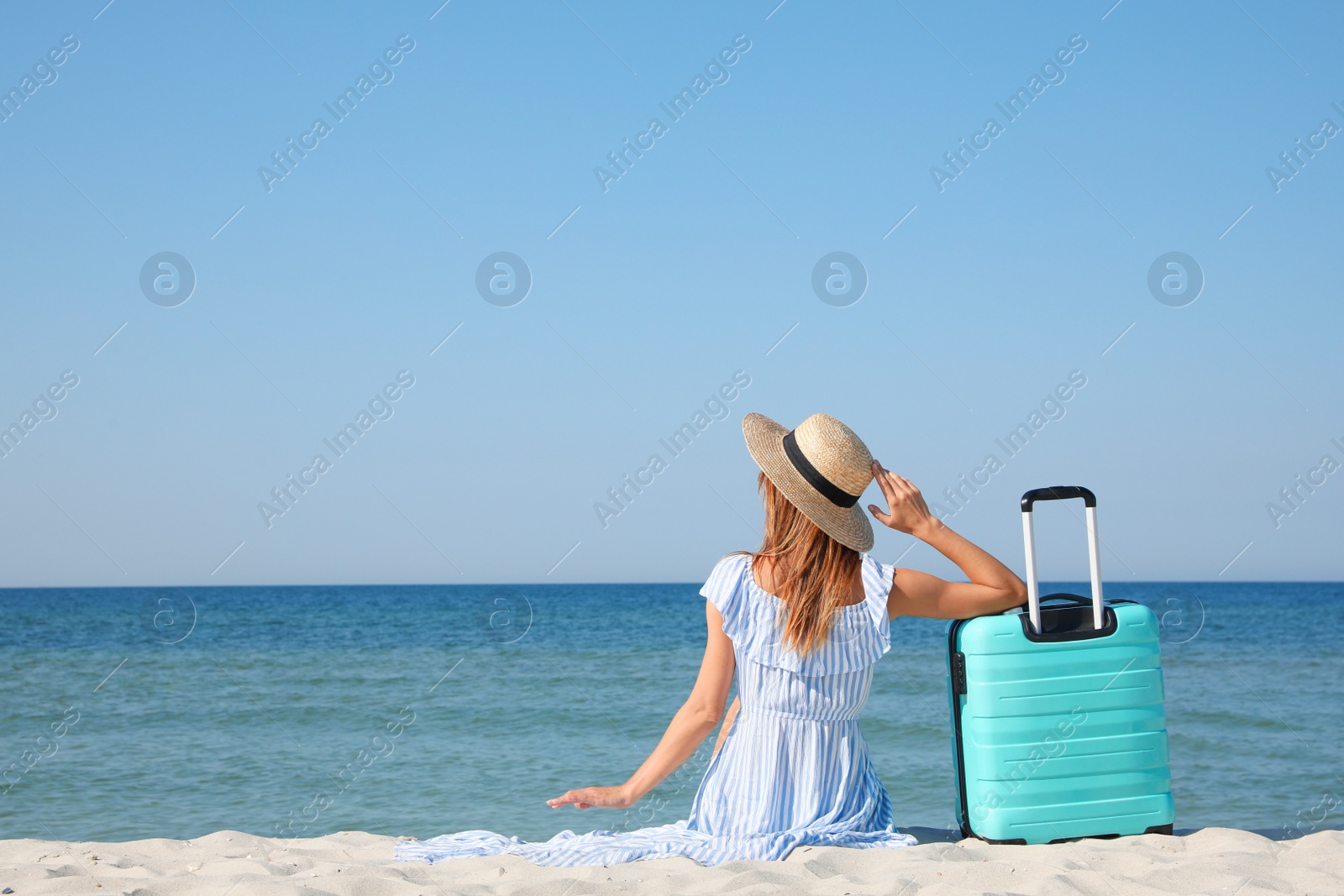 Photo of Woman with suitcase sitting on sandy beach near sea, back view