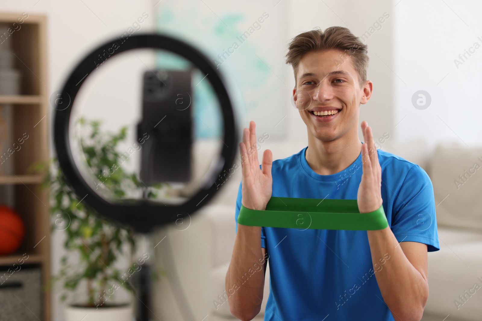 Photo of Smiling sports blogger with resistance band streaming online fitness lesson with smartphone at home