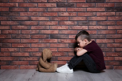 Sad little boy with toy sitting on floor near brick wall. Space for text