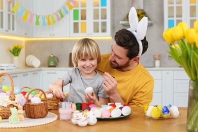Father and his cute son having fun while painting Easter eggs at table in kitchen