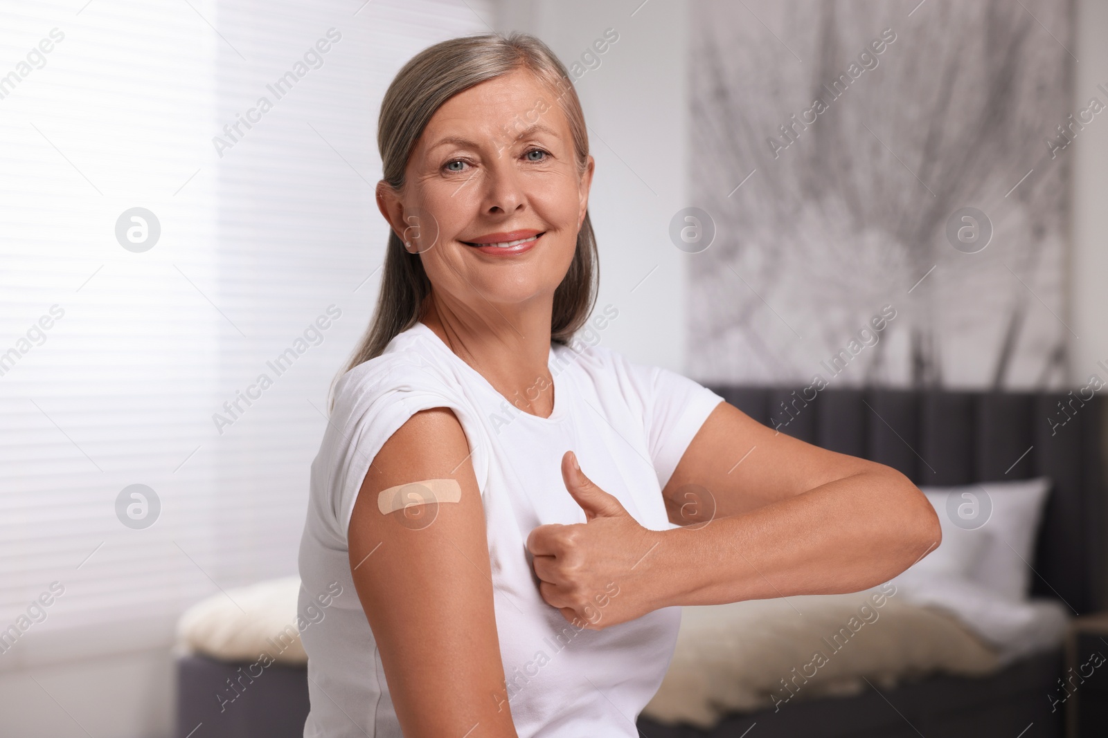 Photo of Senior woman with adhesive bandage on her arm after vaccination showing thumb up indoors