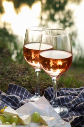 Photo of Glasses of delicious rose wine, cheese and grapes on picnic blanket near lake