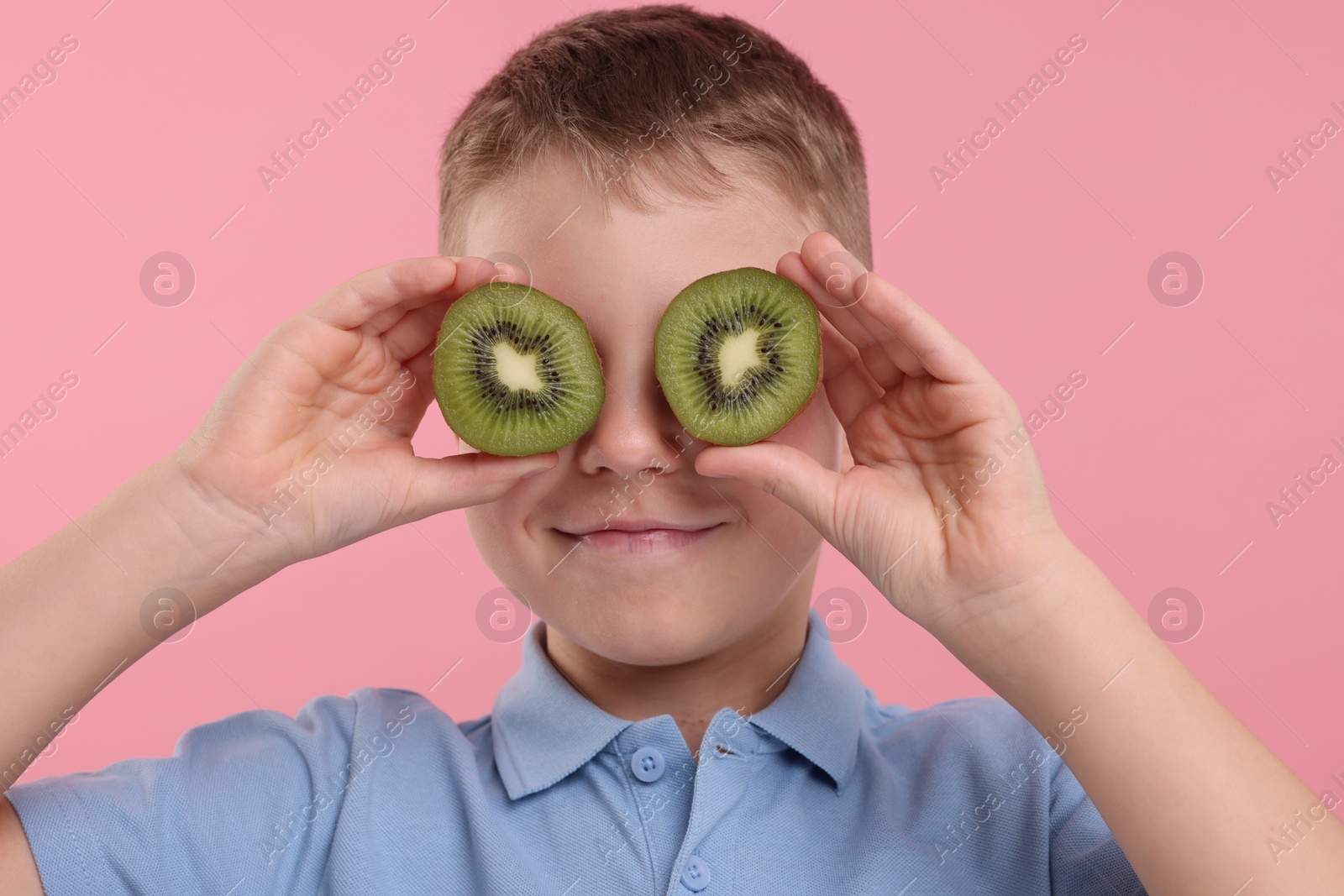 Photo of Boy covering eyes with halves of fresh kiwi on pink background