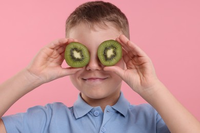 Photo of Boy covering eyes with halves of fresh kiwi on pink background