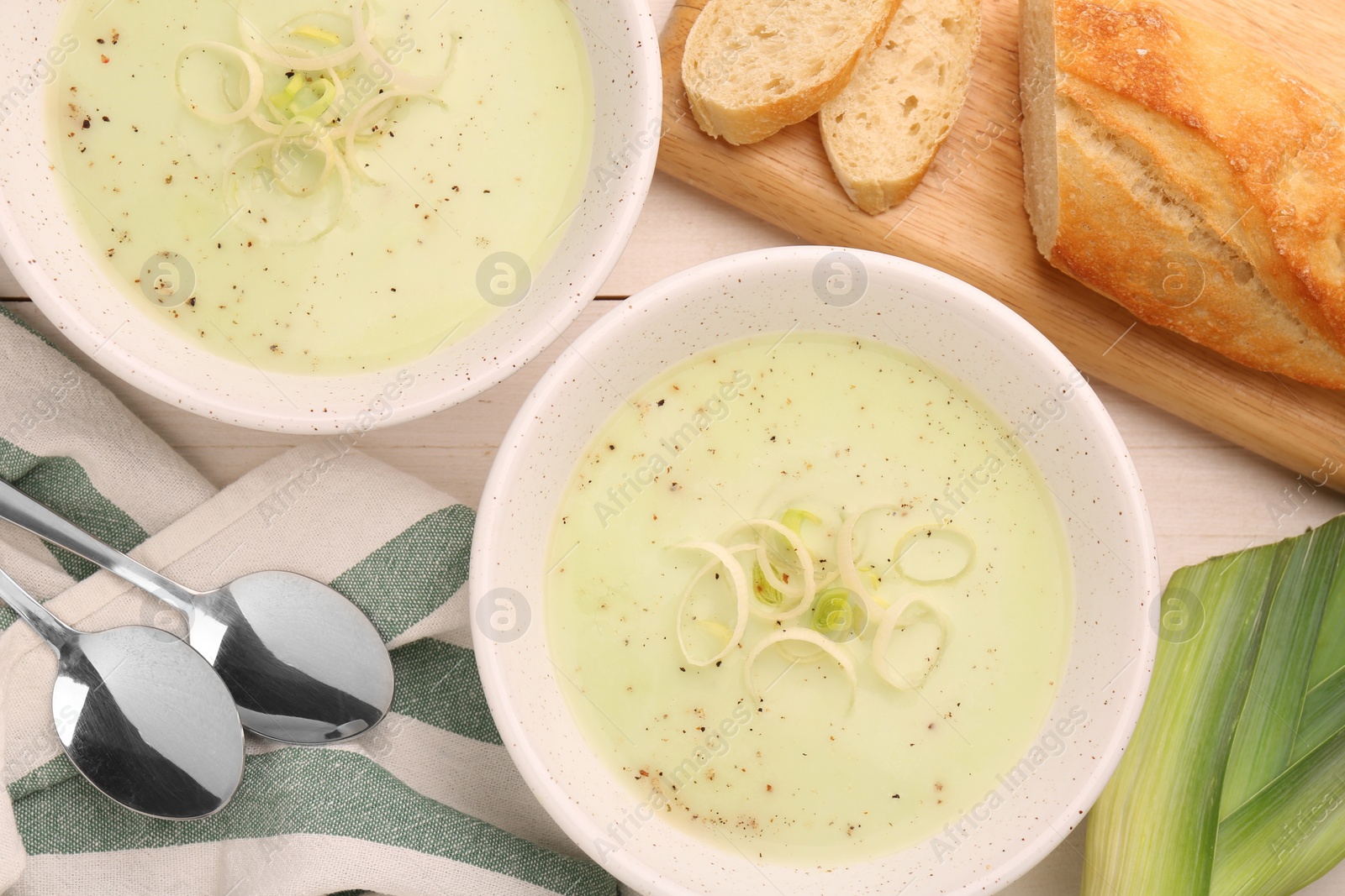 Photo of Bowls of tasty leek soup, spoons and bread on white wooden table, flat lay