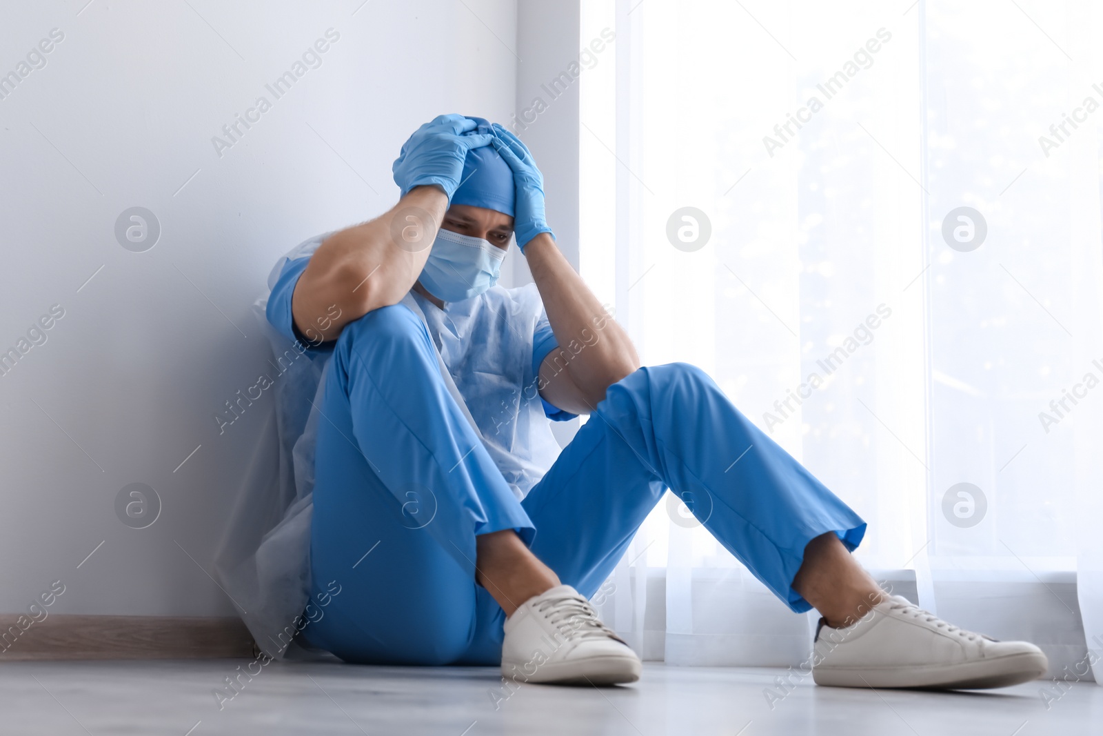 Photo of Exhausted doctor sitting on floor indoors. Stress of health care workers during COVID-19 pandemic