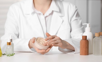 Photo of Dermatologist with jar testing cosmetic product at white table indoors, selective focus