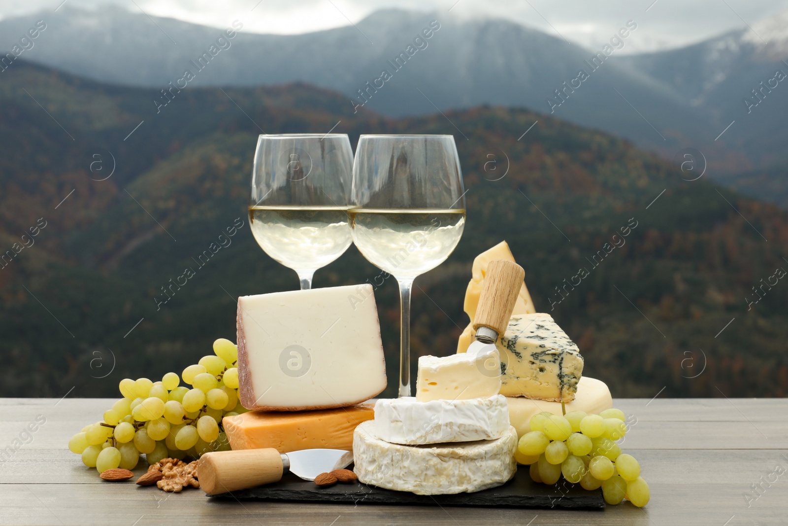 Photo of Different types of delicious cheeses, snacks and wine on wooden table against mountain landscape