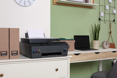 Photo of Modern printer with paper on white chest of drawers near stylish workplace indoors