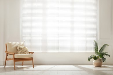 Photo of Soft armchair and houseplant near large window with blinds in spacious room. Interior design
