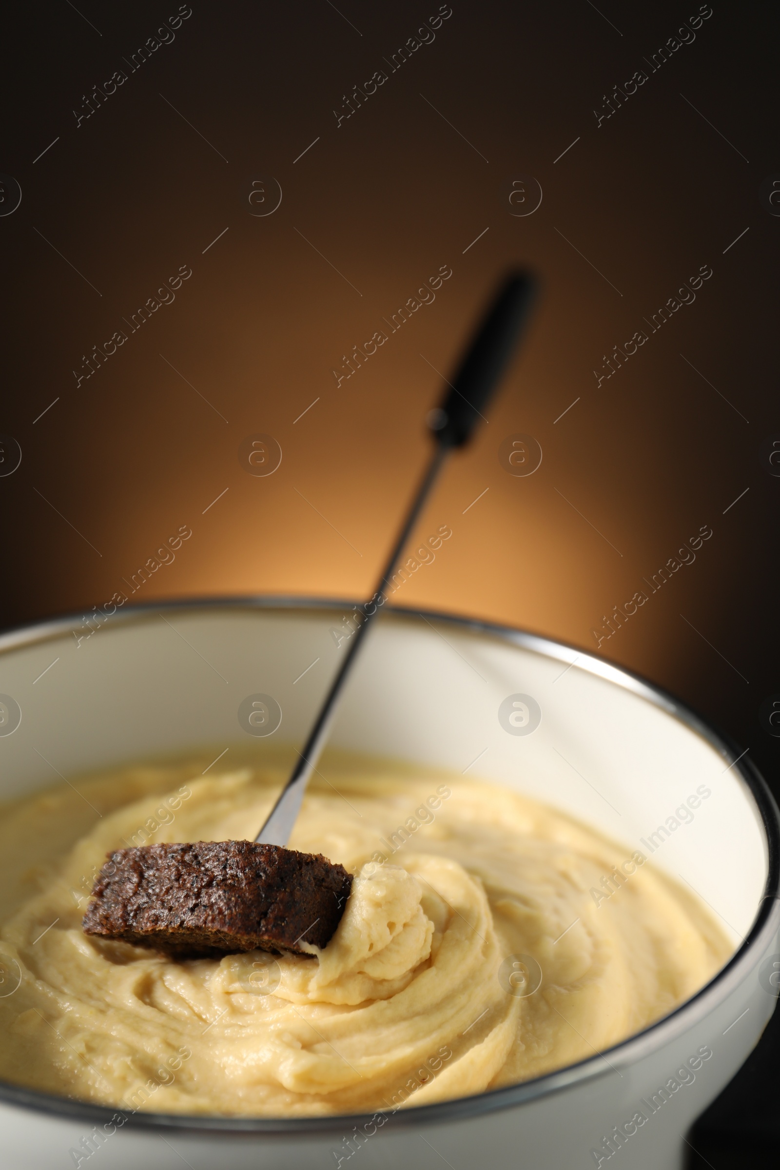 Photo of Fork with piece of bread and melted cheese in fondue pot on dark background, closeup