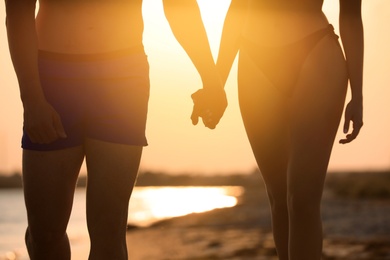 Woman in bikini holding hands with her boyfriend on beach at sunset, closeup. Lovely couple