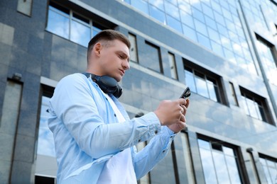 Man with headphones using smartphone near building outdoors, low angle view. Space for text