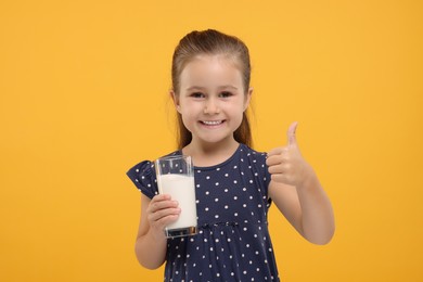 Photo of Cute girl with glass of fresh milk showing thumb up on orange background