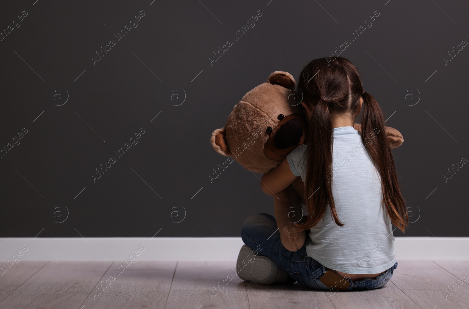 Photo of Child abuse. Upset little girl with teddy bear sitting on floor near gray wall indoors, back view. Space for text