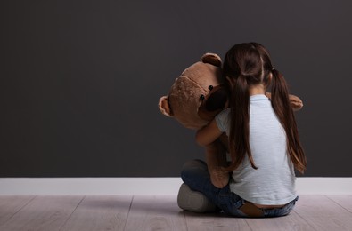 Child abuse. Upset little girl with teddy bear sitting on floor near gray wall indoors, back view. Space for text