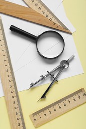 Different rulers, magnifying glass and compass on yellow background, flat lay