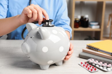 Doctor with stethoscope and piggy bank near pills at wooden table in hospital, closeup. Medical insurance