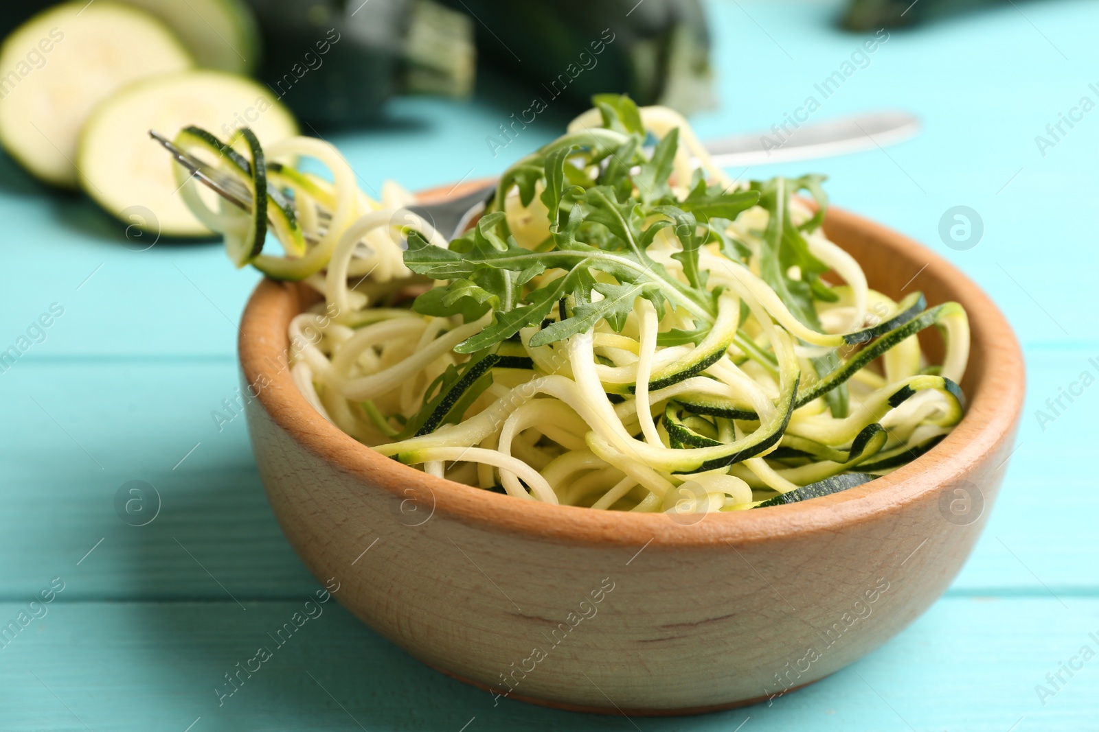 Photo of Delicious zucchini pasta with arugula in bowl on light blue wooden table, closeup