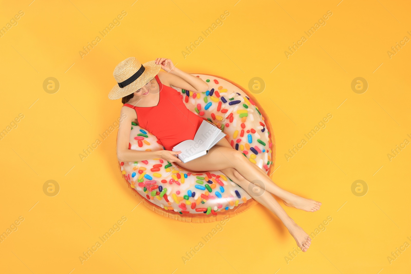 Photo of Happy young woman with beautiful suntan reading book on inflatable ring against orange background, top view