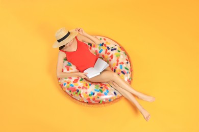 Happy young woman with beautiful suntan reading book on inflatable ring against orange background, top view