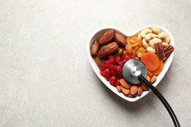 Photo of Heart shaped bowl with dried fruits, nuts and stethoscope on grey background, top view. Space for text