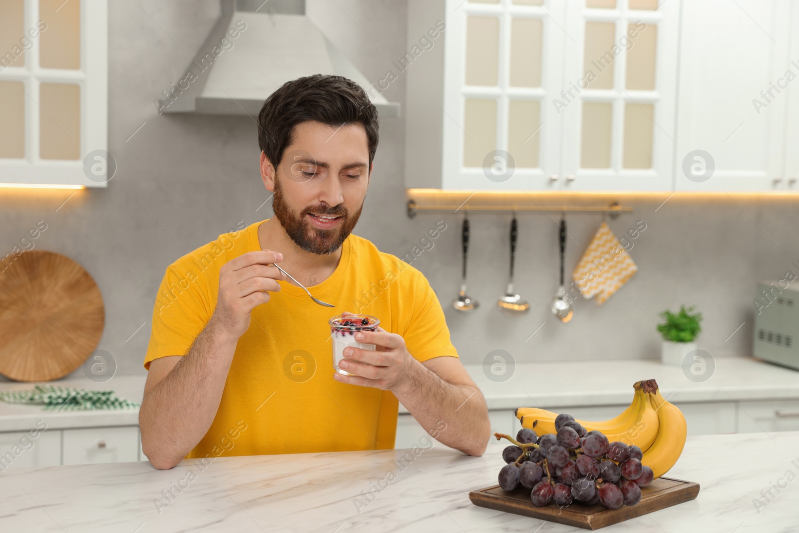 Photo of Handsome man with delicious yogurt at white marble table in kitchen. Space for text