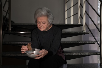 Photo of Poor mature woman with bowl and bread on stairs. Space for text