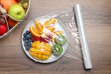 Plate of fresh fruits and berries with plastic food wrap on wooden table, flat lay