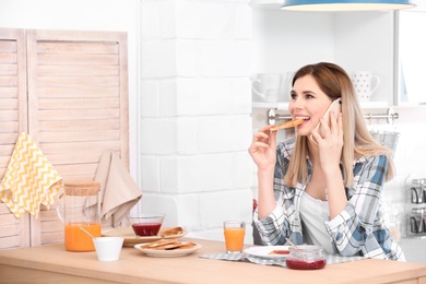 Photo of Beautiful woman eating tasty toasted bread with jam while talking on mobile phone at table