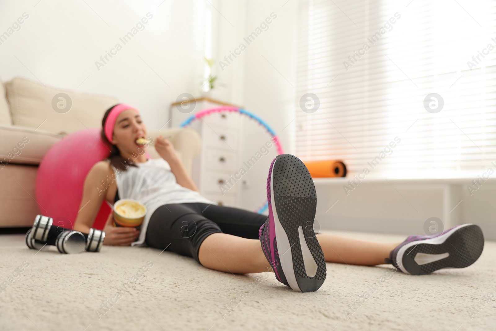 Photo of Lazy young woman eating ice cream instead of training at home, focus on legs