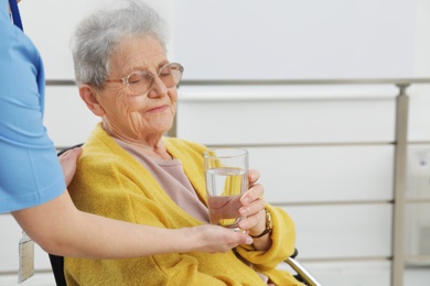 Photo of Nurse giving water to senior woman in wheelchair at hospital. Medical assisting