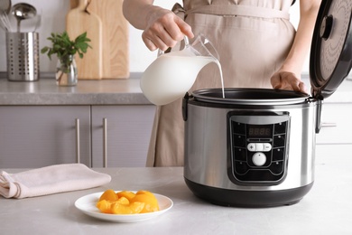 Woman pouring milk into modern multi cooker for making delicious yogurt at table, closeup