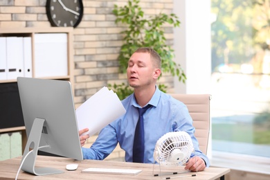 Young man suffering from heat in front of small fan at workplace
