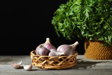 Photo of Fresh raw garlic in wicker basket and parsley on wooden table against black background