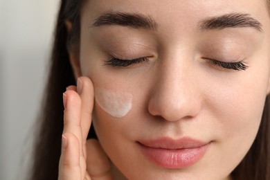 Young woman with dry skin applying cream onto her face on blurred background, closeup